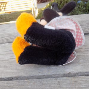 May include: A plush toy with black and orange feet, a black body, and a white and pink polka-dotted skirt. The toy has black ears. The toy is lying on a weathered wooden surface.