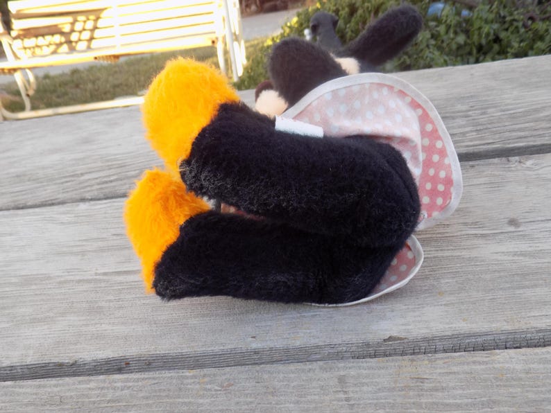 May include: A plush toy with black and orange feet, a black body, and a white and pink polka-dotted skirt. The toy has black ears. The toy is lying on a weathered wooden surface.