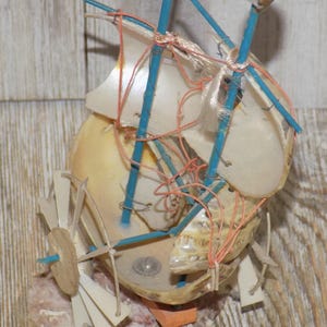 May include: A handcrafted miniature sailboat made from a seashell, featuring blue supports, white sails, and orange and red string details. The boat is set against a wooden background, showcasing a unique nautical theme.