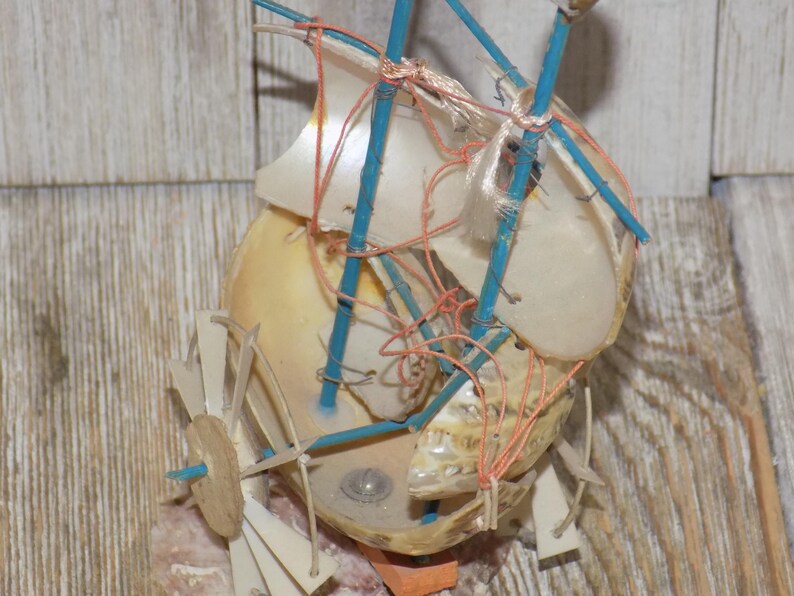 May include: A handcrafted miniature sailboat made from a seashell, featuring blue supports, white sails, and orange and red string details. The boat is set against a wooden background, showcasing a unique nautical theme.