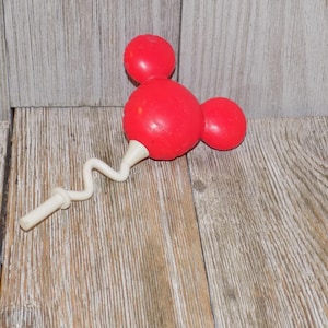 May include: A red plastic toy shaped like Mickey Mouse's head with a white, springy handle. The toy is on a wooden surface.