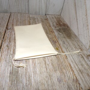 Puede incluir: Una peque&ntilde;a bolsa de cord&oacute;n de color blanco roto hecha de tela con un cord&oacute;n blanco. La bolsa est&aacute; sobre una superficie de madera.