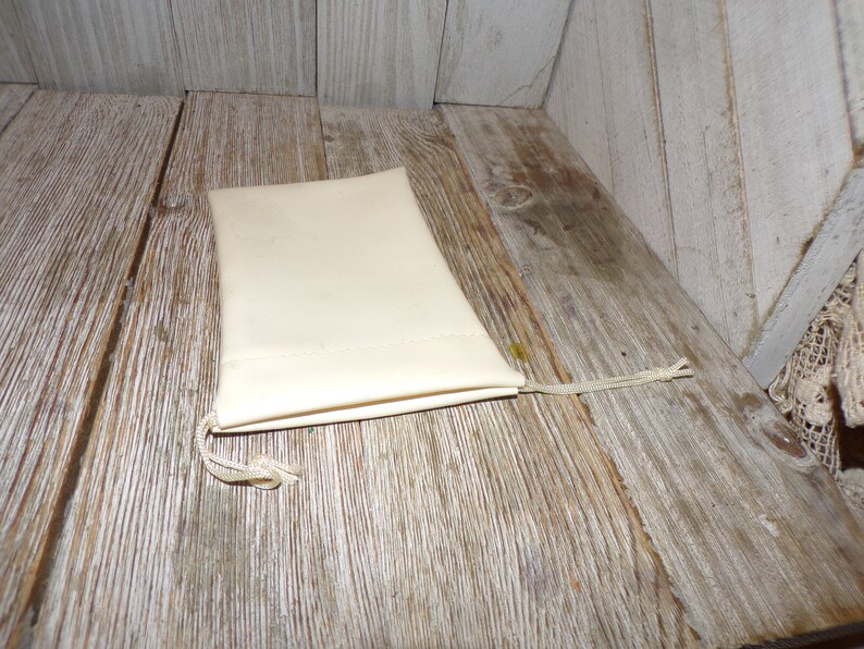 Puede incluir: Una peque&ntilde;a bolsa de cord&oacute;n de color blanco roto hecha de tela con un cord&oacute;n blanco. La bolsa est&aacute; sobre una superficie de madera.