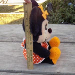 May include: A plush Minnie Mouse doll, featuring black fur, a white face, and a red and white polka dot dress. The doll has a yellow bow and orange accents. The doll measures approximately 28 cm in height.