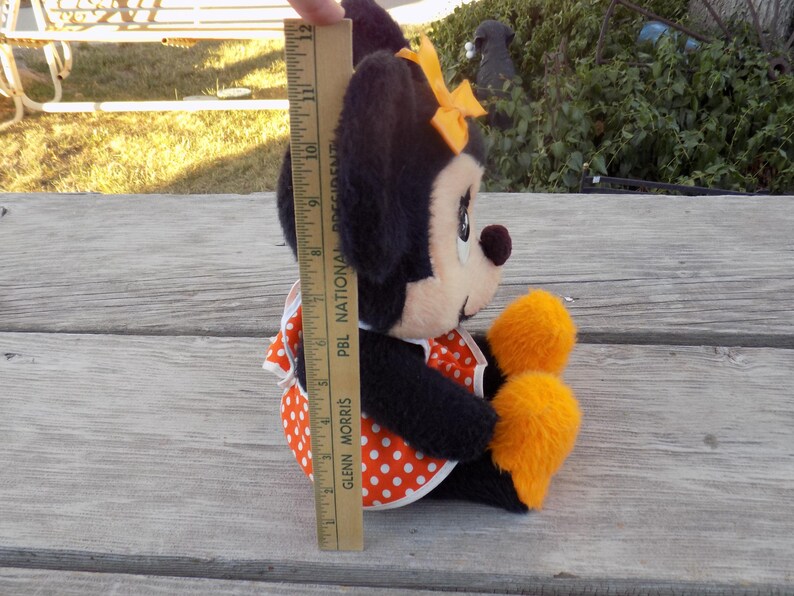 May include: A plush Minnie Mouse doll, featuring black fur, a white face, and a red and white polka dot dress. The doll has a yellow bow and orange accents. The doll measures approximately 28 cm in height.