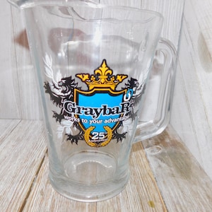 May include: Clear glass pitcher with a handle. The pitcher features a logo with a blue shield, gold crown, and the text "Graybar" and "Works to your advantage." The pitcher also has a "25th Anniversary" emblem.