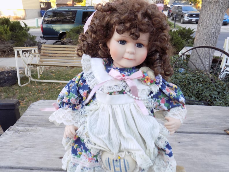 May include: A porcelain doll with brown curly hair and blue eyes. The doll wears a blue floral dress with a white lace collar and apron. A pink ribbon and pearl necklace adorn the doll's neck. The doll is sitting on a wooden surface.