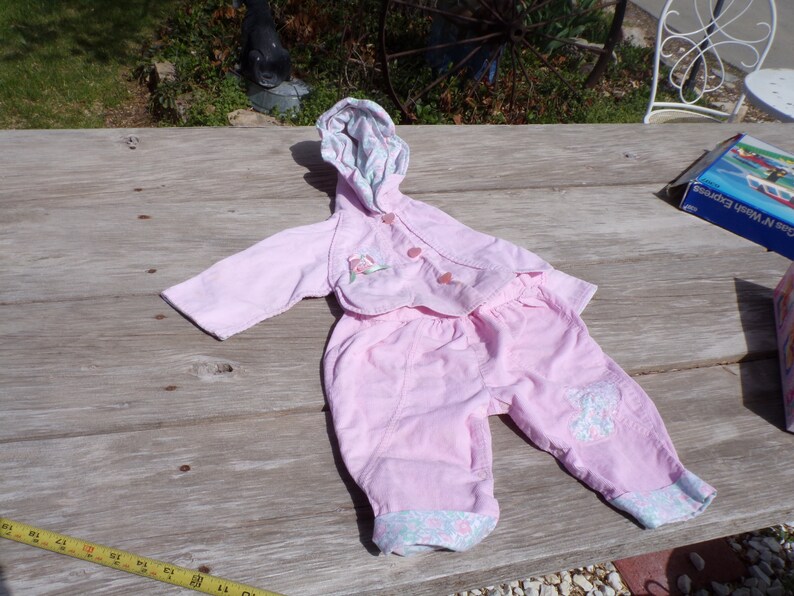 Puede incluir: Un conjunto de beb&eacute; rosa con una chaqueta con capucha y pantalones. La chaqueta tiene un dise&ntilde;o floral y los pantalones tienen un dise&ntilde;o de mariposa.