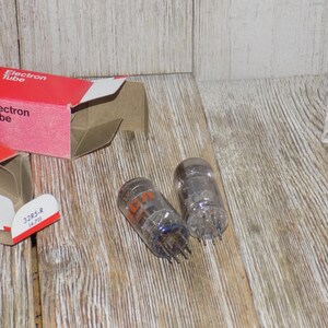 May include: Two clear glass RCA Electron Tubes with orange lettering and metal pins, next to a red and white RCA Electron Tube box. The box is open, revealing the tubes. The tubes are on a wooden surface.
