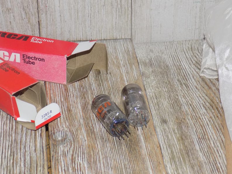May include: Two clear glass RCA Electron Tubes with orange lettering and metal pins, next to a red and white RCA Electron Tube box. The box is open, revealing the tubes. The tubes are on a wooden surface.