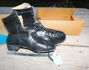 May include: Pair of vintage black ice skates with laces, metal blades, and a tag. The skates are in a box. The boots are leather and have a high-top design. The blades are silver-colored.