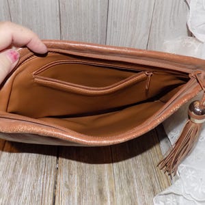May include: A brown leather clutch purse with a zipper closure and a tassel. The purse has a small interior pocket.