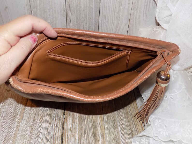 May include: A brown leather clutch purse with a zipper closure and a tassel. The purse has a small interior pocket.