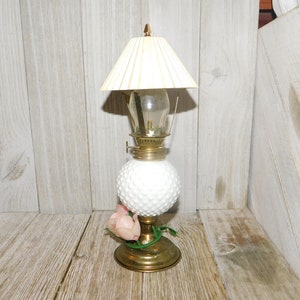 May include: A white milk glass oil lamp with a white pleated lampshade and a single pink flower accent. The lamp has a gold base and a clear glass chimney.