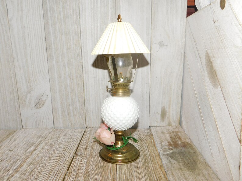 May include: A white milk glass oil lamp with a white pleated lampshade and a single pink flower accent. The lamp has a gold base and a clear glass chimney.