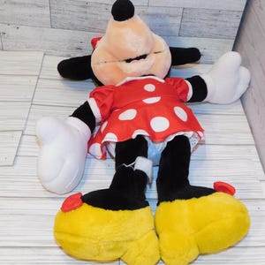 Op de afbeelding: Een pluche Minnie Mouse-pop, met een rode jurk met witte stippen, zwarte armen en benen en grote gele schoenen. De pop heeft witte handschoenen, een rode strik en een lachend gezicht. Het speelgoed ligt op een lichtgekleurd houten oppervlak.