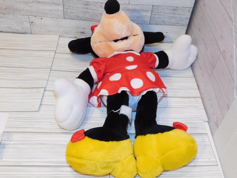 Op de afbeelding: Een pluche Minnie Mouse-pop, met een rode jurk met witte stippen, zwarte armen en benen en grote gele schoenen. De pop heeft witte handschoenen, een rode strik en een lachend gezicht. Het speelgoed ligt op een lichtgekleurd houten oppervlak.