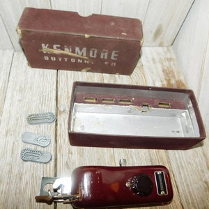 May include: Vintage Kenmore buttonhole sewing machine in a burgundy red color with a black knob and a silver plate with the brand name. The machine comes with a box and three different sized buttonhole attachments.
