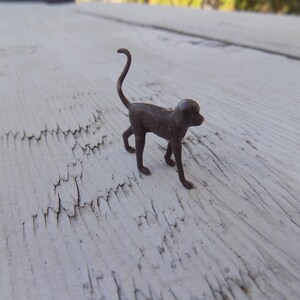 May include: A small, dark brown metal figurine of a monkey standing on all fours. The monkey has a slender, curved tail pointing upward, a rounded head, and four distinct legs. The surface of the metal appears textured and aged with a matte finish.