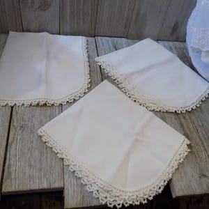 May include: Three white handkerchiefs with crocheted lace edges are arranged on a weathered wooden surface. The handkerchiefs are square with rounded corners. The lace trim adds a delicate, vintage aesthetic.