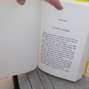 May include: An open book with a yellow cover and white pages. The text on the page reads "Chapter 1 A Curious Stranger". The text continues with a story about Nancy Drew and her friends.