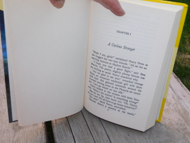 May include: An open book with a yellow cover and white pages. The text on the page reads "Chapter 1 A Curious Stranger". The text continues with a story about Nancy Drew and her friends.