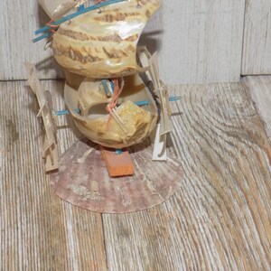 May include: A handcrafted seashell sculpture resembling a boat. The sculpture is made from multiple seashells, with wooden supports and orange and blue accents. The base is a large, pinkish-brown shell, and the structure is set against a wooden background.