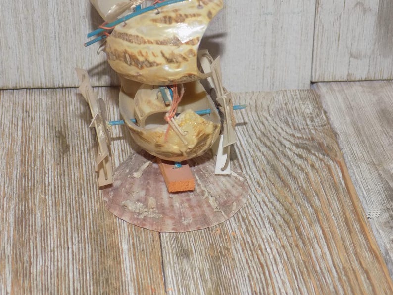 May include: A handcrafted seashell sculpture resembling a boat. The sculpture is made from multiple seashells, with wooden supports and orange and blue accents. The base is a large, pinkish-brown shell, and the structure is set against a wooden background.