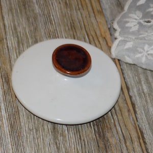 May include: A white ceramic lid with a brown knob. The lid is round and sits on a wooden surface. A white lace fabric is in the background.