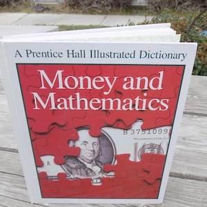 May include: A Prentice Hall Illustrated Dictionary book titled "Money and Mathematics." The cover features a red puzzle design revealing a portion of a U.S. dollar bill. The book is open, showing the pages.