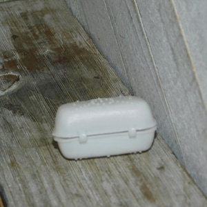 Op de afbeelding: Een kleine, witte plastic container met een scharnierend deksel en een getextureerde bovenkant. De container is rechthoekig met afgeronde randen en ligt op een verweerd houten oppervlak.