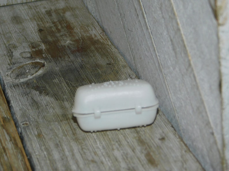 Op de afbeelding: Een kleine, witte plastic container met een scharnierend deksel en een getextureerde bovenkant. De container is rechthoekig met afgeronde randen en ligt op een verweerd houten oppervlak.