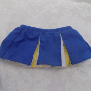May include: A blue pleated skirt featuring white and yellow fabric inserts between the pleats. The skirt has an elasticized waistband and a short, flared silhouette, designed with a layered appearance showing contrasting colors.