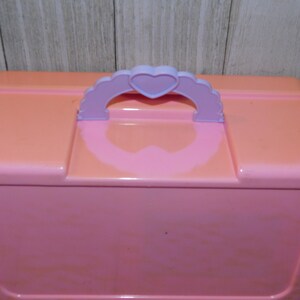 Puede incluir: Una caja de almacenamiento de pl&aacute;stico rosa con un asa en forma de coraz&oacute;n morado. La caja tiene un acabado liso y brillante.