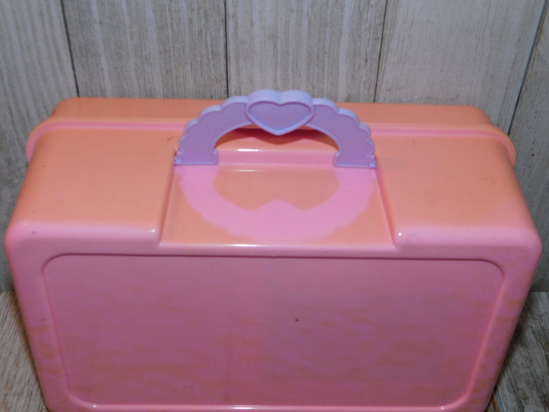 Puede incluir: Una caja de almacenamiento de pl&aacute;stico rosa con un asa en forma de coraz&oacute;n morado. La caja tiene un acabado liso y brillante.
