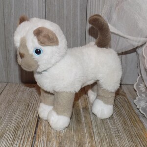 May include: A plush toy cat with white fur and tan markings on its ears, face, and legs. The cat has blue eyes and a curled-up tail. It is standing on a wooden surface.