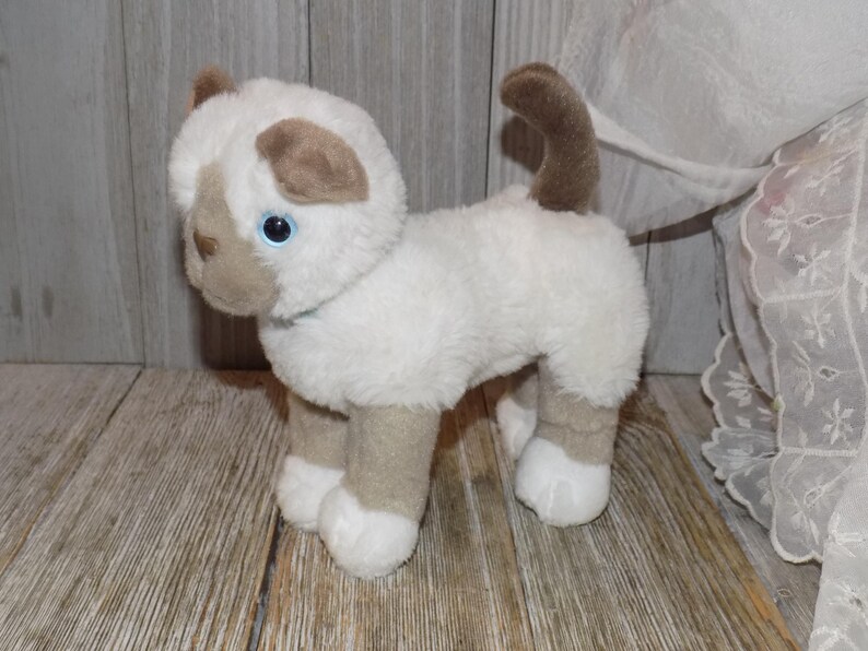 May include: A plush toy cat with white fur and tan markings on its ears, face, and legs. The cat has blue eyes and a curled-up tail. It is standing on a wooden surface.