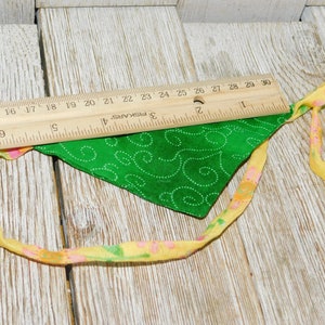 May include: A green fabric bandana with a white swirl pattern and yellow and pink trim. The bandana is lying on a wooden surface with a ruler.