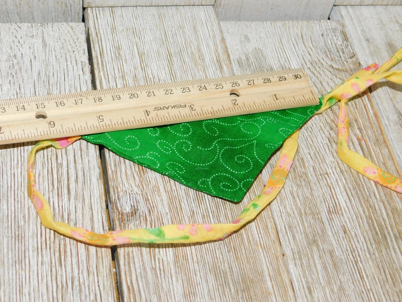May include: A green fabric bandana with a white swirl pattern and yellow and pink trim. The bandana is lying on a wooden surface with a ruler.