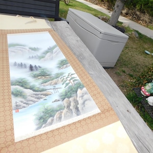 May include: A landscape painting on a scroll, depicting a misty mountain scene with a river, trees, and a small bridge. The artwork features a brown border with a repeating circular pattern. The scroll is laid out on a wooden surface.