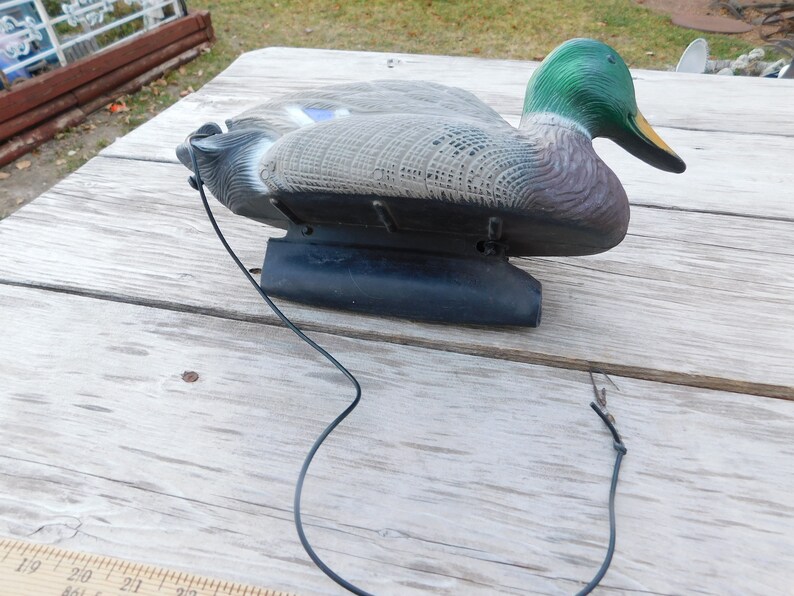 May include: A green-headed mallard duck decoy with a black base and a string attached. The decoy is made of plastic and has a realistic appearance.