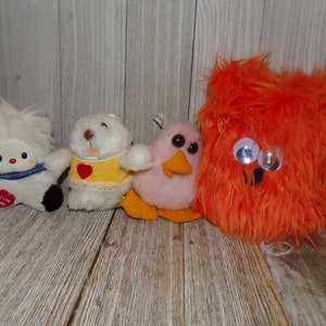 May include: Four plush toys, a white fluffy one with a blue and white scarf and a heart that says "I love you", a white one with a yellow dress and a red heart, a pink bird with orange feet and a yellow beak, and a bright orange fuzzy one with large googly eyes.
