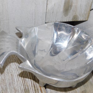 May include: A silver-colored, fish-shaped bowl with a textured surface. The bowl has a wide, open design with a tail and fins, and rests on three small feet. The bowl is made of metal.