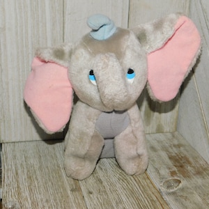 May include: A gray plush toy elephant with pink ears and a blue hat. The elephant has blue eyes and a gray trunk.