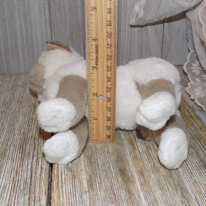 May include: A plush toy cat in a lying position, with white and tan fur. The toy is approximately 5 inches long, as measured by a wooden ruler. The cat's paws and ears are tan, and the body is white.