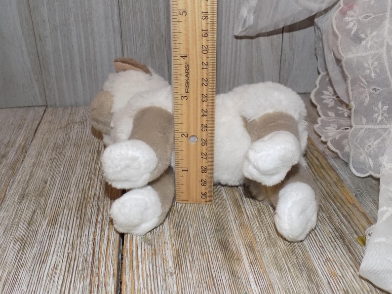 May include: A plush toy cat in a lying position, with white and tan fur. The toy is approximately 5 inches long, as measured by a wooden ruler. The cat's paws and ears are tan, and the body is white.
