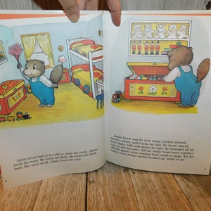 May include: A children's book illustration featuring a brown bear with a blue shirt and overalls. The bear is standing in front of a red toy chest filled with toys. The text on the page reads, "Jamie knew exactly how many stuffed animals, blocks, robots, and trucks he had. He knew exactly how many balls and games he had. He arranged all his toys in special ways. All the teddy bears were together. All the games were stacked from small to large. All the trains were always neatly hooked up, ready to go."