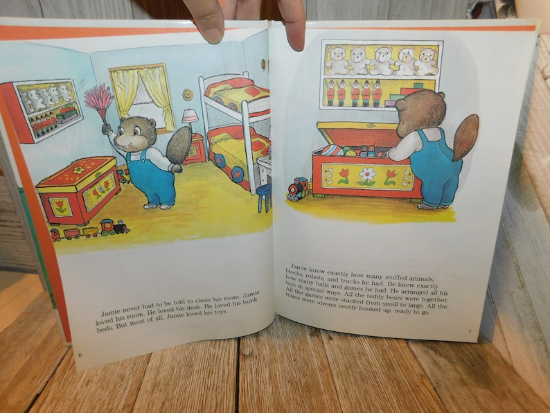 May include: A children's book illustration featuring a brown bear with a blue shirt and overalls. The bear is standing in front of a red toy chest filled with toys. The text on the page reads, "Jamie knew exactly how many stuffed animals, blocks, robots, and trucks he had. He knew exactly how many balls and games he had. He arranged all his toys in special ways. All the teddy bears were together. All the games were stacked from small to large. All the trains were always neatly hooked up, ready to go."