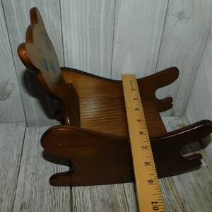 Puede incluir: Una silla mecedora de madera con forma de oso con la cara pintada. La silla es marr&oacute;n y tiene una regla al lado para referencia de tama&ntilde;o. La silla mide aproximadamente 25 cm de largo.