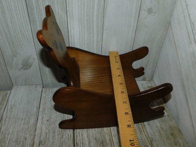Puede incluir: Una silla mecedora de madera con forma de oso con la cara pintada. La silla es marr&oacute;n y tiene una regla al lado para referencia de tama&ntilde;o. La silla mide aproximadamente 25 cm de largo.